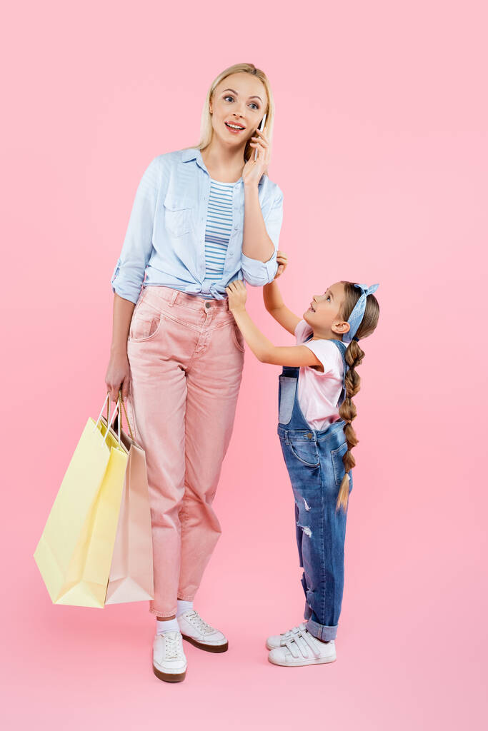 longitud completa de la madre feliz sosteniendo bolsas de compras y hablando en el teléfono inteligente cerca de la hija en rosa  - Foto, imagen
