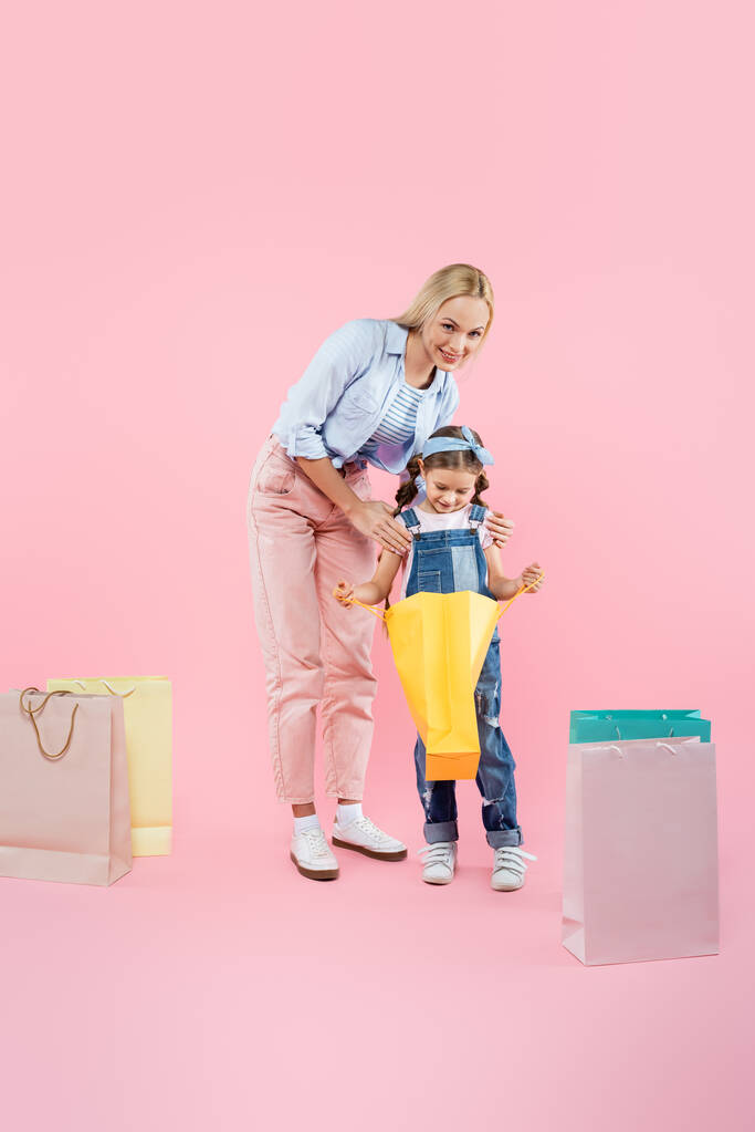 pleine longueur de mère heureuse et fille souriante regardant sac à provisions sur rose - Photo, image