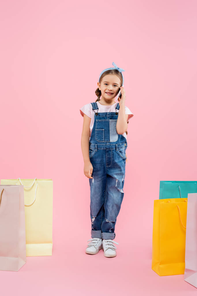 comprimento total da criança feliz falando no smartphone perto de sacos de compras em rosa - Foto, Imagem