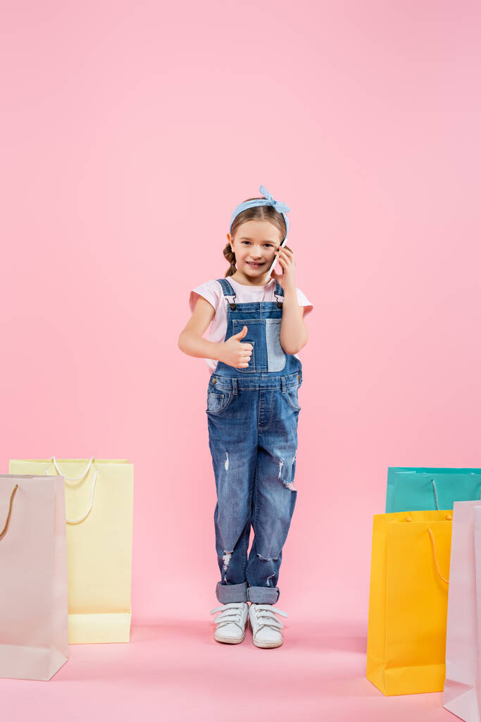 полный рост ребенка, говорящего по смартфону и показывающего большой палец рядом с сумками с покупками на розовом фоне - Фото, изображение