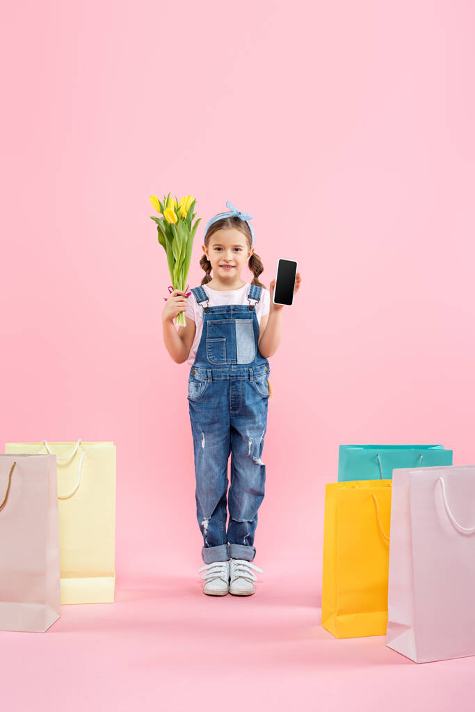 longitud completa de niño sosteniendo teléfono inteligente con pantalla en blanco y tulipanes cerca de bolsas de compras en rosa - Foto, Imagen