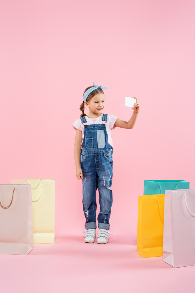 longitud completa de niño alegre tomar selfie en el teléfono inteligente cerca de bolsas de compras en rosa - Foto, imagen