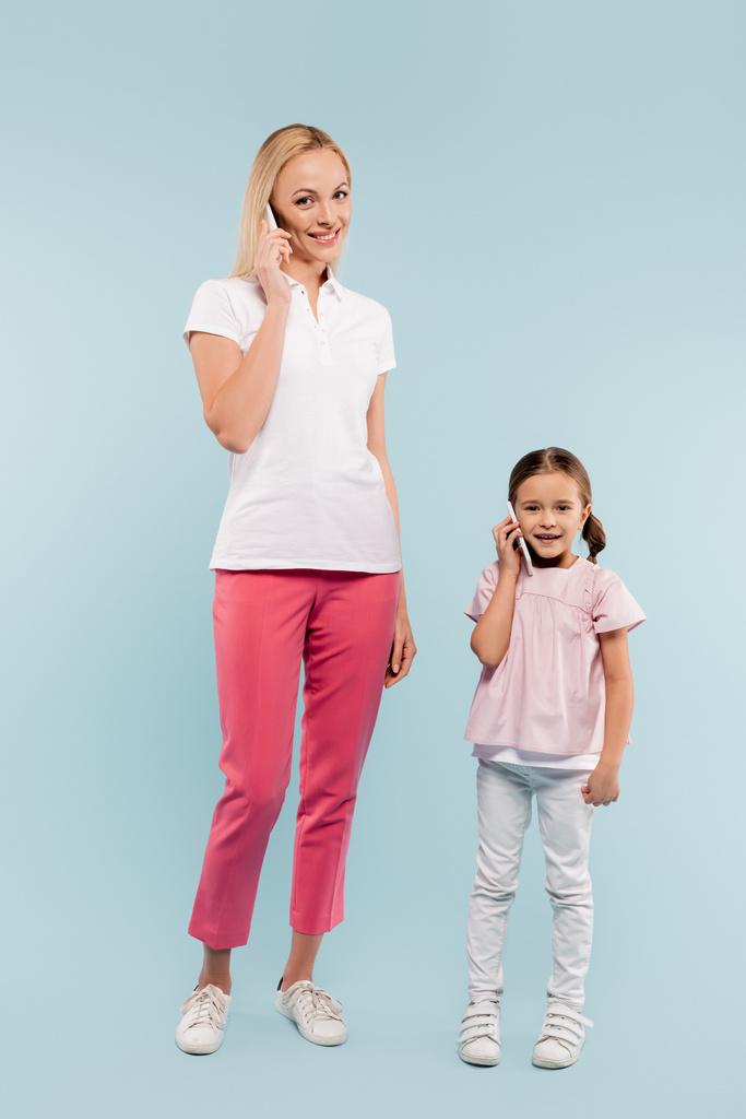 full length of cheerful mother and daughter talking on smartphones on blue - Photo, Image