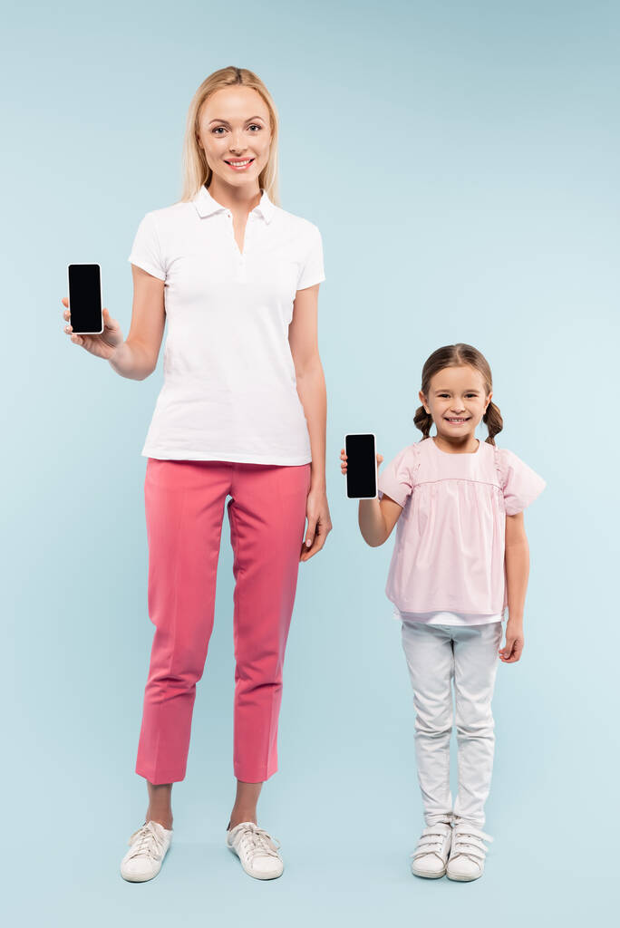 full length of cheerful mother and daughter holding smartphones with blank screen on blue - Photo, Image