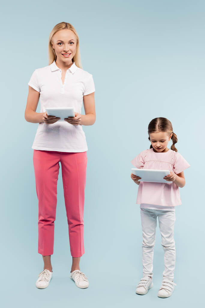 longitud completa de alegre madre e hija sosteniendo tabletas digitales en azul - Foto, Imagen