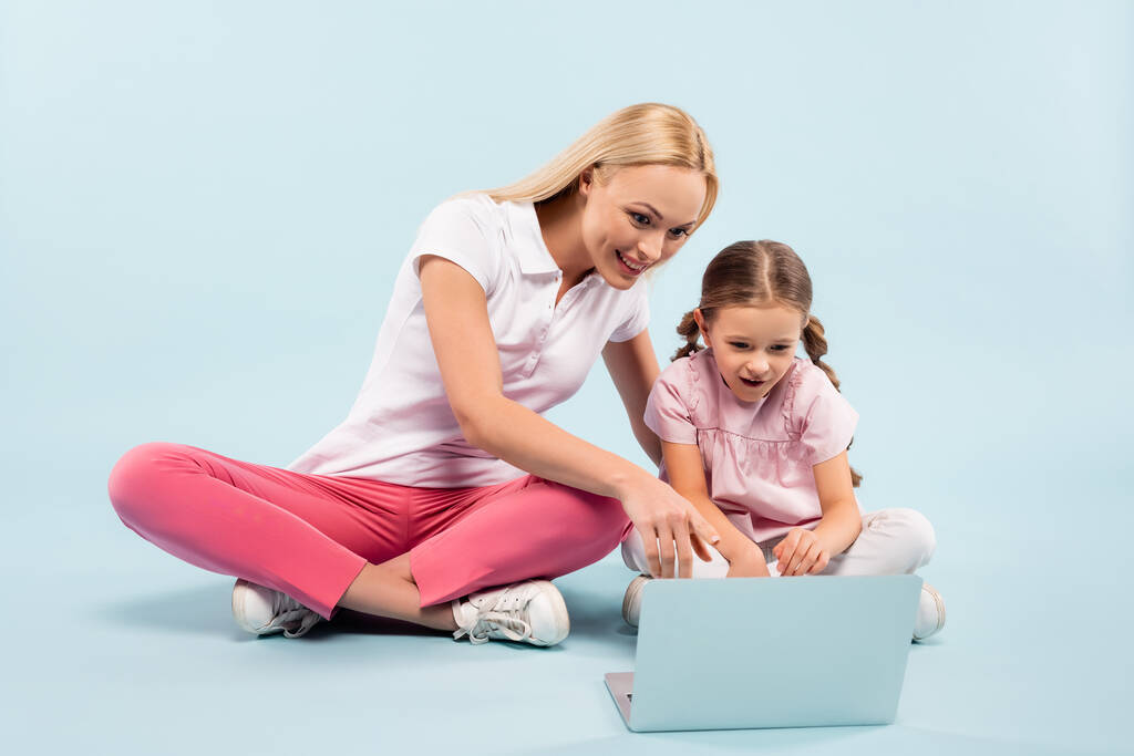 sorridente mãe e filha sentado com pernas cruzadas e apontando com laptop dedo no azul - Foto, Imagem