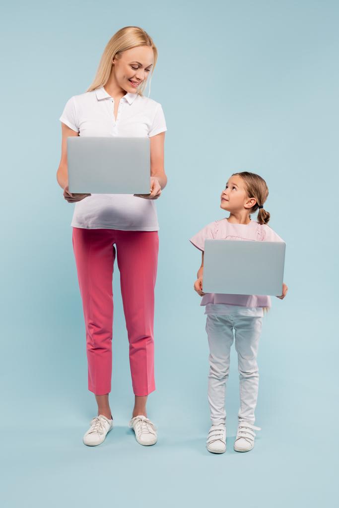 full length of mother and daughter standing with laptops and looking at each other on blue - Photo, Image