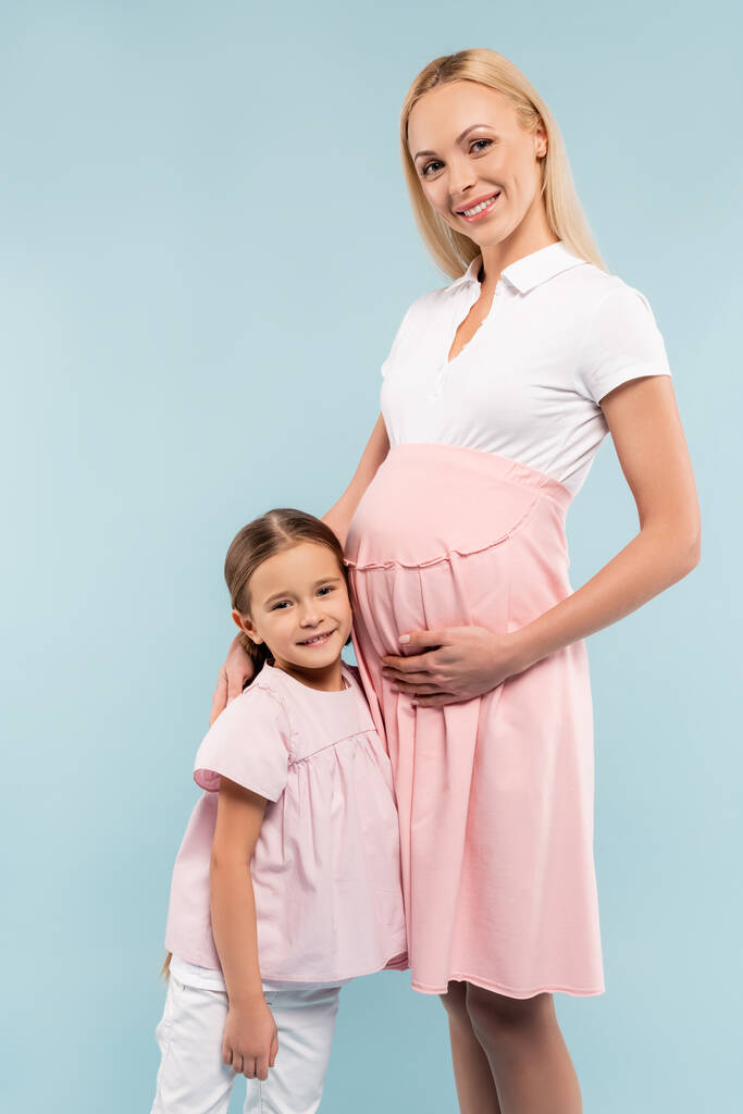 cheerful daughter standing with pregnant mother isolated on blue - Photo, Image