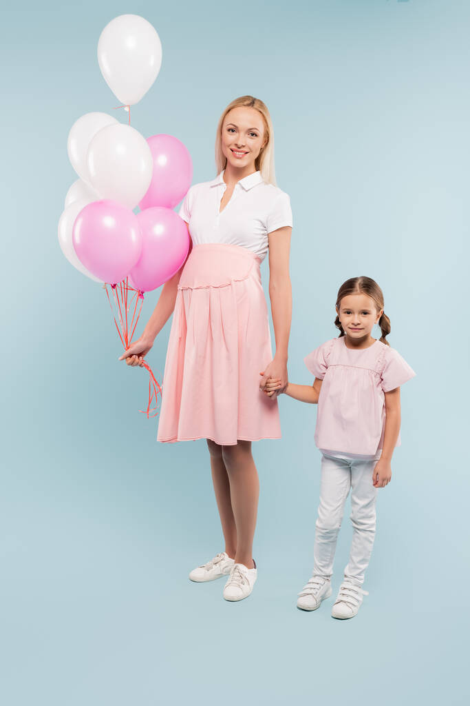 volle Länge der glücklichen schwangeren Frau hält Luftballons in der Nähe der Tochter auf blau - Foto, Bild