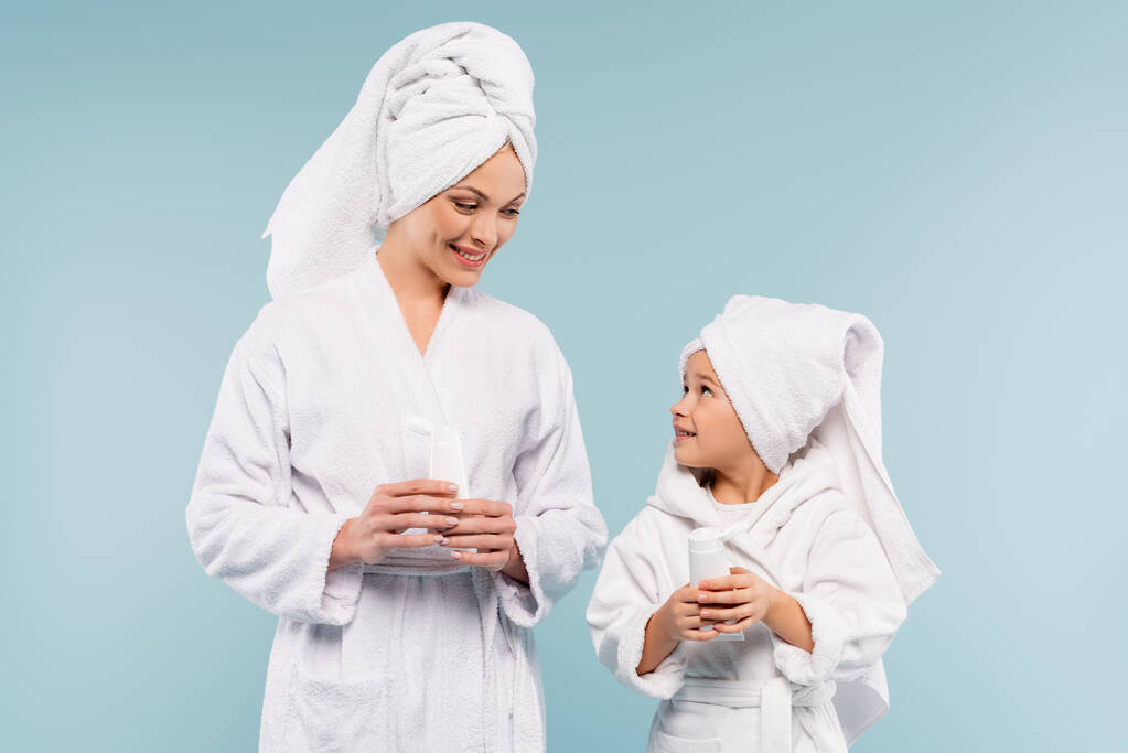 feliz madre e hijo en batas de baño sosteniendo tubos con crema cosmética aislada en azul - Foto, Imagen