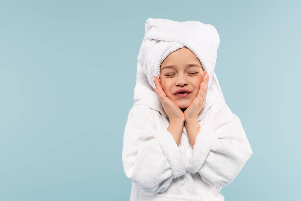 enfant heureux en peignoir et serviette sur la tête souriant tout en touchant les joues isolé sur bleu - Photo, image