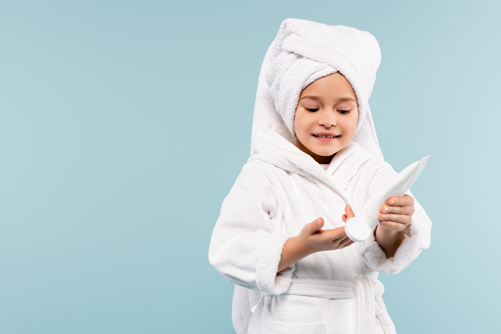 lachend kind in badjas en handdoek op hoofdbeugel met cosmetisch product geïsoleerd op blauw - Foto, afbeelding