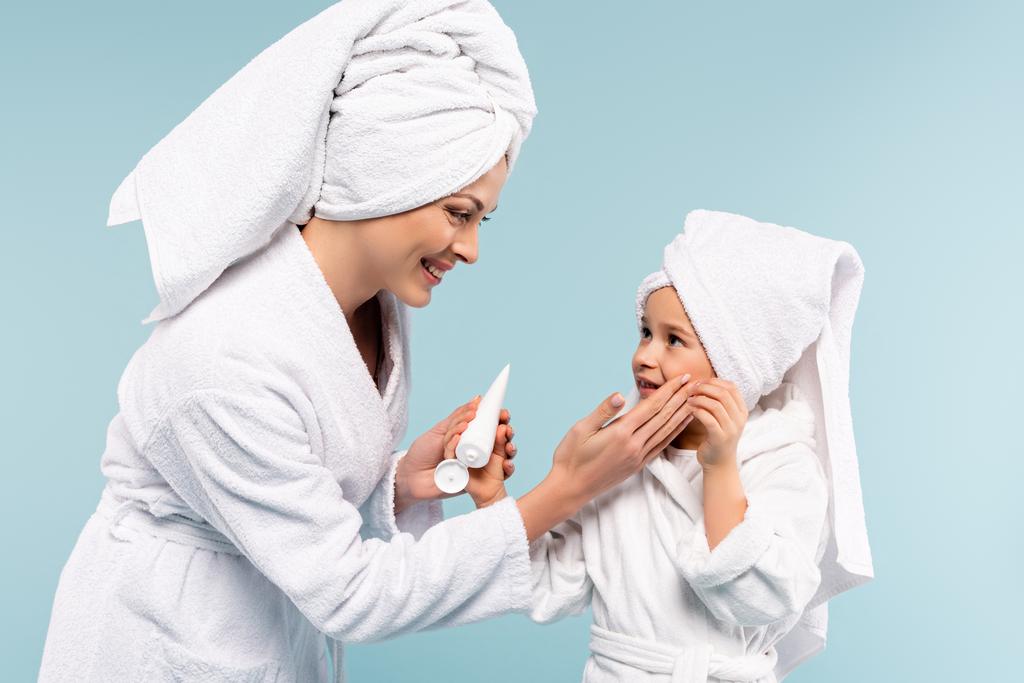 mãe feliz em roupão de banho segurando tubo e aplicando creme cosmético no rosto da filha isolada no azul - Foto, Imagem