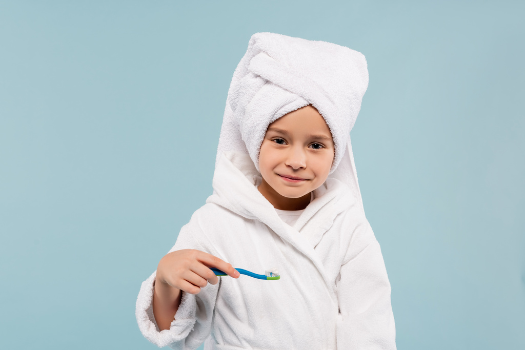 glückliches Kind im Bademantel und Handtuch auf dem Kopf, Zahnbürste mit Zahnpasta isoliert auf blau - Foto, Bild