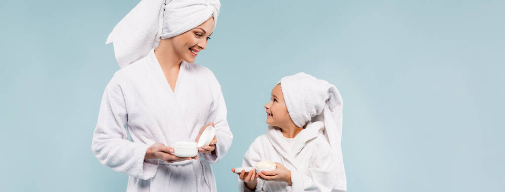 Bornozlu mutlu anne ve çocuk mavi pankartta izole edilmiş kozmetik krem kapları tutuyorlar. - Fotoğraf, Görsel