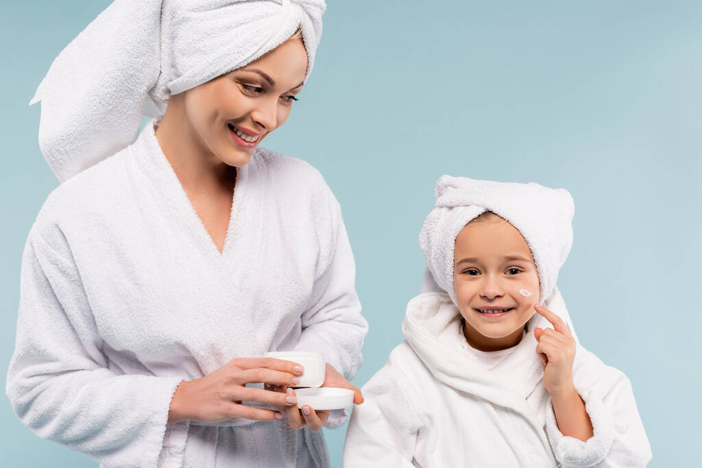 glückliche Mutter im Bademantel mit Behälter und Blick auf Tochter, die Kosmetikcreme auf Gesicht aufträgt, isoliert auf blauem Grund - Foto, Bild