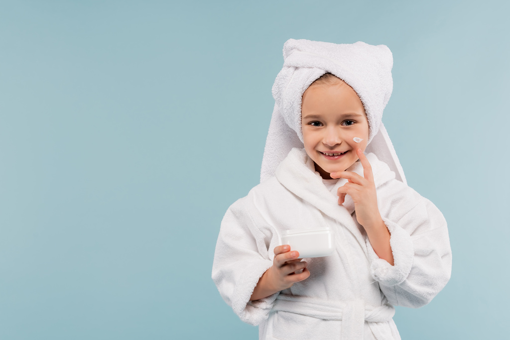 cheerful child in bathrobe and towel on head holding container and applying cosmetic product isolated on blue - Photo, Image