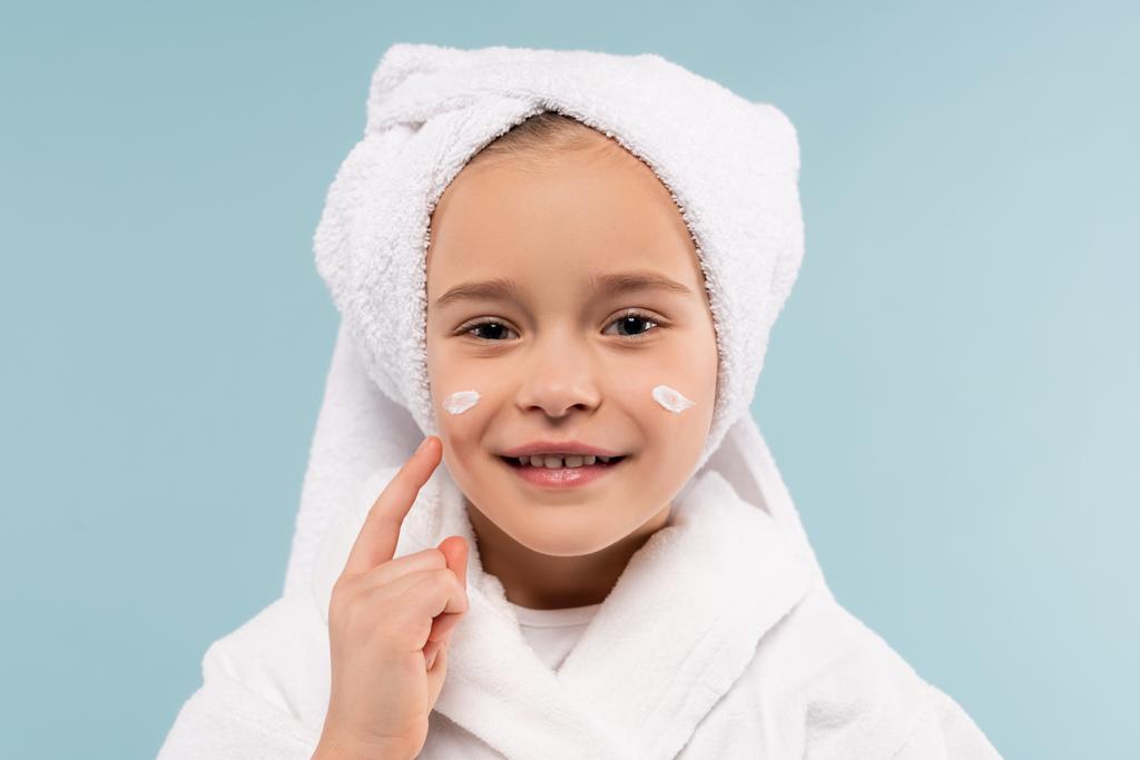 fröhliches Kind in Bademantel und Handtuch auf dem Kopf, kosmetische Creme isoliert auf blau auftragen - Foto, Bild