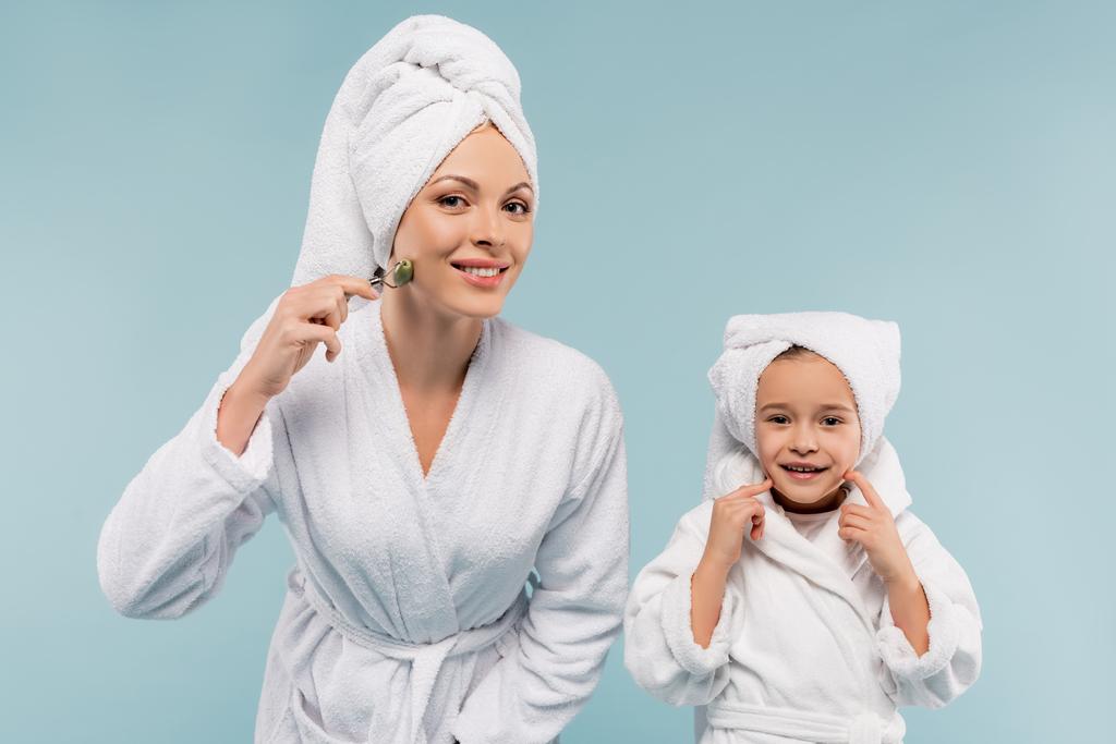glückliches Kind im Bademantel lächelt nahe Mutter mit Jade-Rolle isoliert auf blau - Foto, Bild