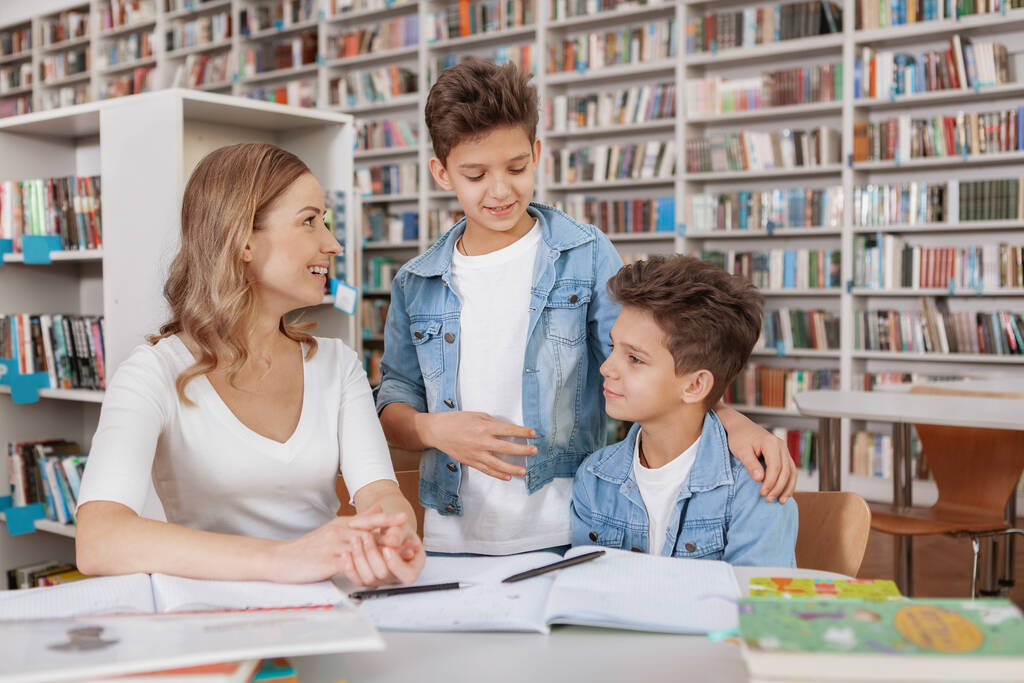 Два брата-близнеца и их мама наслаждаются совместными занятиями в библиотеке. Красивая женщина помогает своим детям с домашним заданием - Фото, изображение