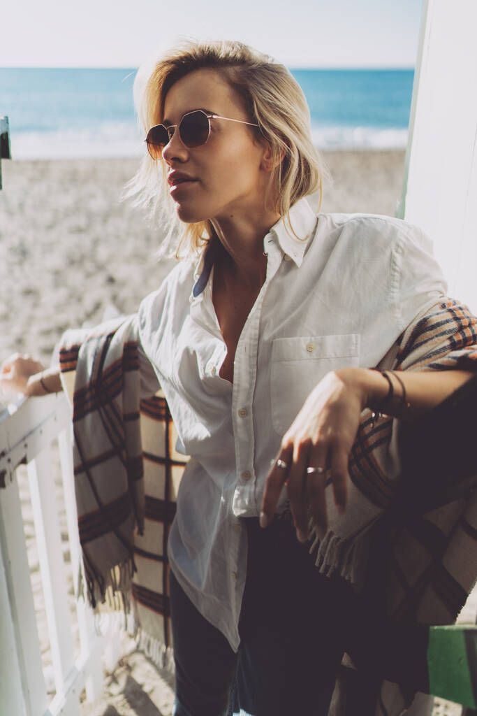 Charmante kaukasische Frau mit langen blonden Haaren und moderner Sonnenbrille mit weißem Hemd und blauer Jeans schaut zur Seite, während sie mit der Decke auf den Schultern am Sandstrand steht.. - Foto, Bild