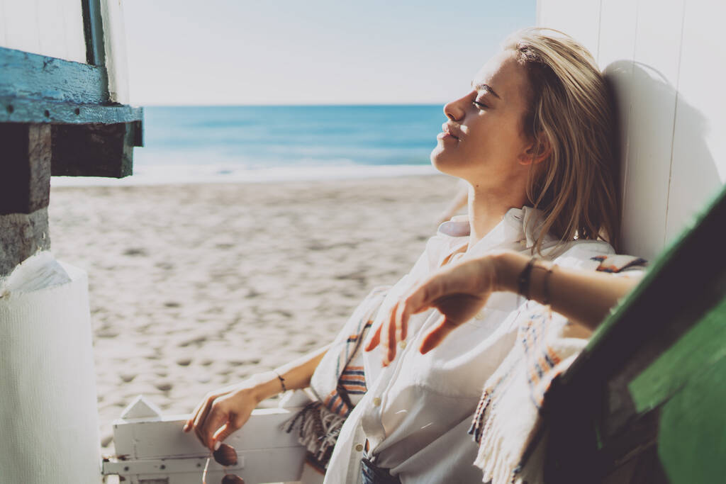 Retrato vista lateral de una mujer rubia caucásica con los ojos cerrados usando camisa blanca de pie apoyada en la pared de madera de una casa de playa mientras disfruta del calor del sol sobre un fondo borroso lado del mar. - Foto, Imagen