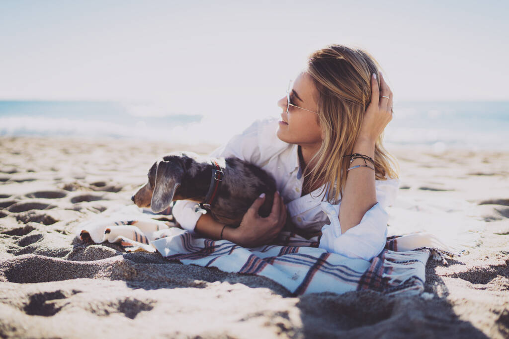 Glückliche kaukasische Mädchen, die Spaß mit Hund genießen Freizeit zum Spazierengehen und Training. Unbekümmerte Frau im weißen Hemd, die sich auf einer Decke am Meer ausruht, nachdem sie mit einem Welpen am Strand spaziert ist - Foto, Bild