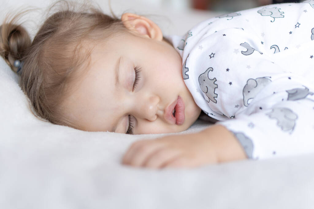 子供時代、ケア、母親、健康、医学、小児科の概念-閉じる小さな平和穏やかな幼児の就学前の女の子は、柔らかいベッドの上でおなかの上に寝そべって深い昼寝をします。就寝時のmod閉じる目 - 写真・画像