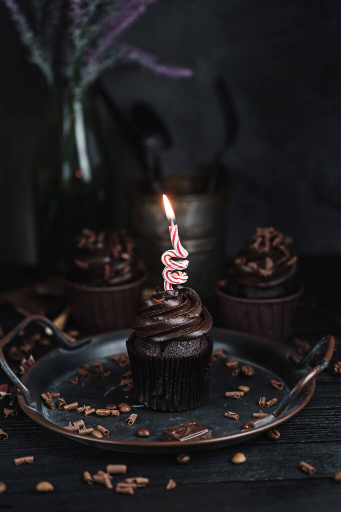 黒テーブルでチョコレート型のクリームといくつかのマフィンやカップケーキ。チョコレートケーキの上にお祝いのろうそくが燃える - 写真・画像