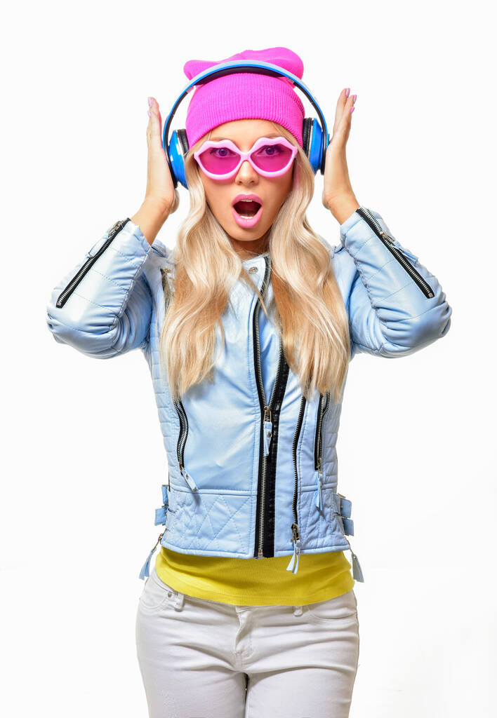 Moda dość beztroska kobieta słuchanie muzyki w słuchawkach w kolorowy różowy kapelusz, niebieska kurtka i okulary przeciwsłoneczne. - Zdjęcie, obraz