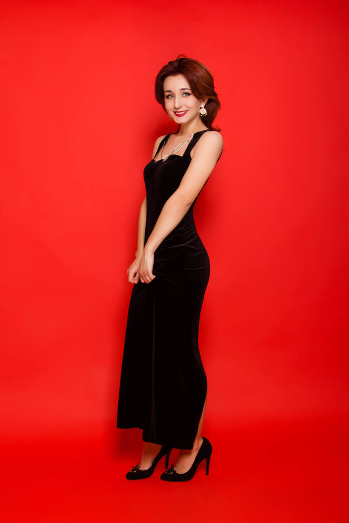 Piękna, młoda kobieta w czarnej sukience i czarnych butach pozujących na czerwonym tle. - Zdjęcie, obraz