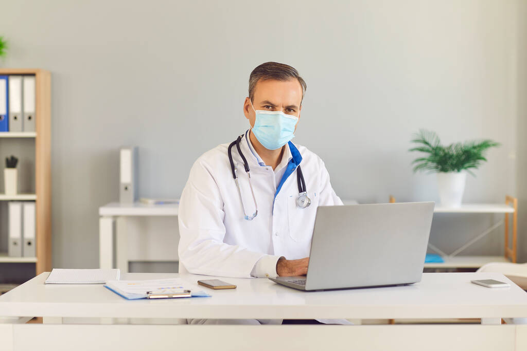 Σοβαρός γιατρός με προστατευτική μάσκα κάθεται στο γραφείο του με ένα φορητό υπολογιστή και κοιτάζοντας την κάμερα. - Φωτογραφία, εικόνα