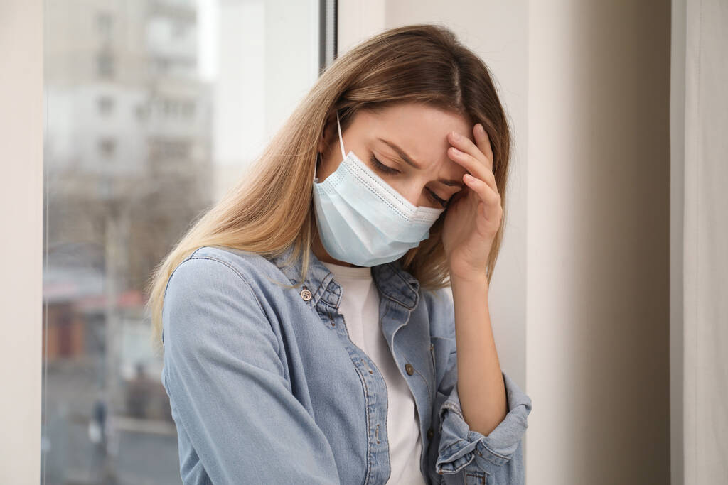 Traurige Frau in Schutzmaske neben Fenster drinnen. Während der Coronavirus-Pandemie zu Hause bleiben - Foto, Bild
