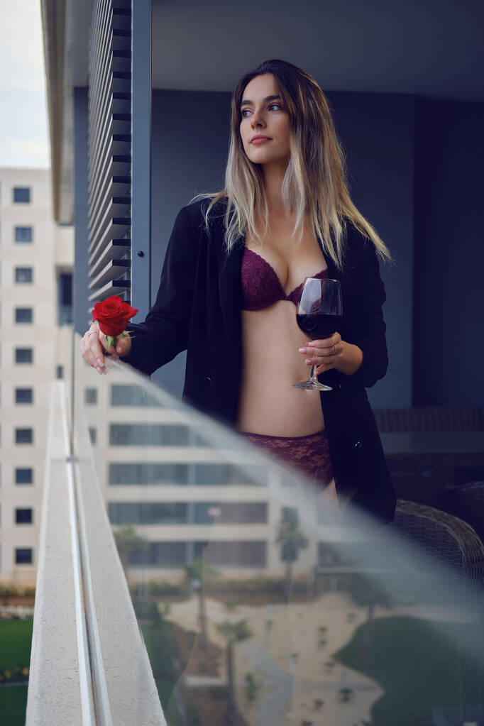 Mladá krásná žena nosí spodní prádlo bundu stojí sám na balkoně drží červenou růži a sklenici vína dívá do dálky. Módní a kosmetický koncept - Fotografie, Obrázek