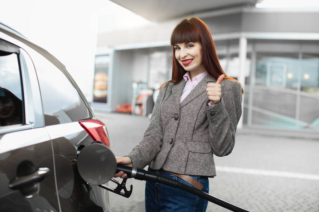 Attractive cheerful young business woman, smiling and showing her thumb up to camera, while refueling her luxury car at gas station - Photo, Image