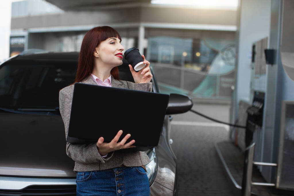 Geschäftsfrau an der Tankstelle. Hübsche junge Frau benutzt ihren Laptop und trinkt Kaffee to go, während sie ihr Auto an der Tankstelle tankt - Foto, Bild