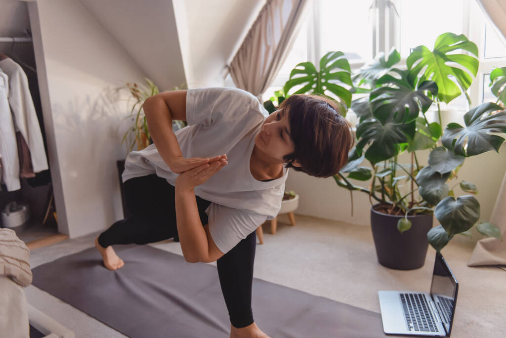 Una mujer en una alfombra haciendo giro y estirando la posición asana en su dormitorio. Práctica de yoga en línea en casa. Un estilo de vida saludable. Entrenamiento en línea en casa durante un bloqueo. Enfoque selectivo suave. - Foto, imagen