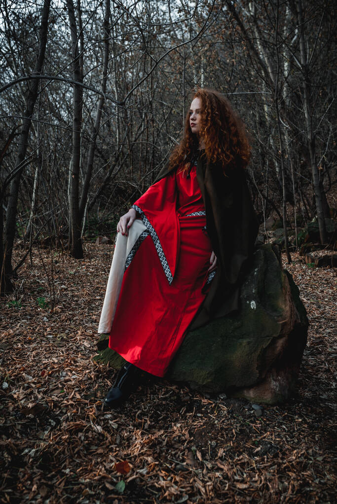 Vörös hajú nő piros ruhában, történelmi kelta jelmezben az őszi erdőben. - Fotó, kép