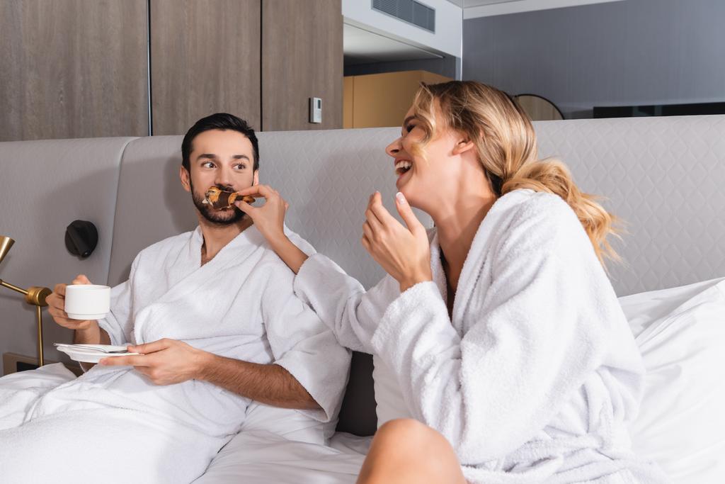 Femme gaie nourrissant croissant au petit ami musulman avec café sur le lit de l'hôtel  - Photo, image