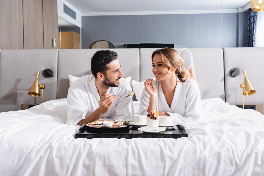 Smiling interracial couple looking at each other near breakfast on tray on hotel bed  - Photo, Image