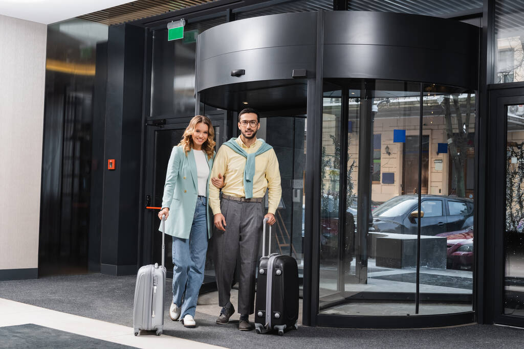 Веселая межрасовая пара с чемоданами, стоящими у дверей отеля  - Фото, изображение