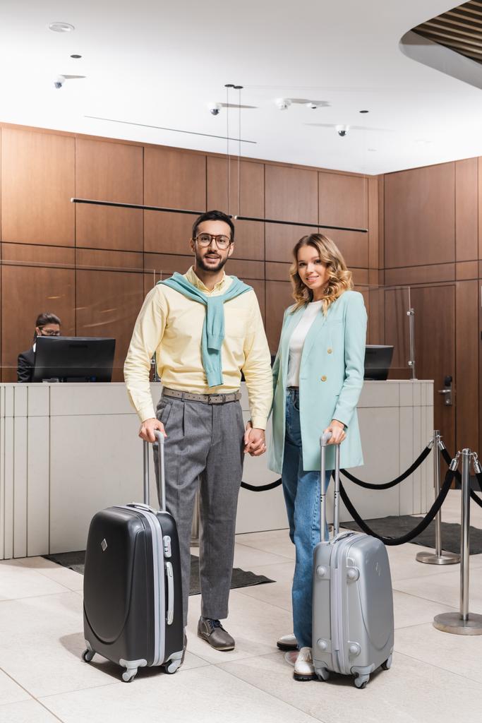 Χαμογελώντας διαφυλετικό ζευγάρι με βαλίτσες κρατώντας τα χέρια στο λόμπι του ξενοδοχείου  - Φωτογραφία, εικόνα
