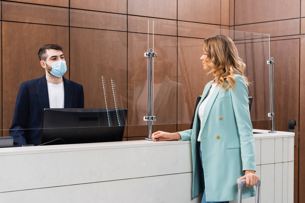 Recepční v lékařské masce při pohledu na ženu s kufrem v hotelové hale  - Fotografie, Obrázek