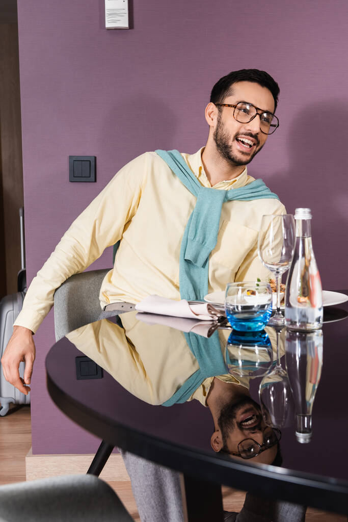 Fröhlicher muslimischer Mann sitzt neben köstlichem Abendessen im Hotelzimmer  - Foto, Bild