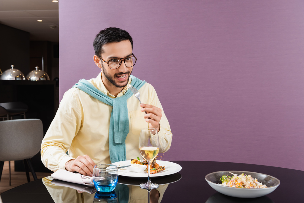 Hombre árabe sonriente sosteniendo tenedor cerca de la comida y el vino en la habitación del hotel  - Foto, imagen