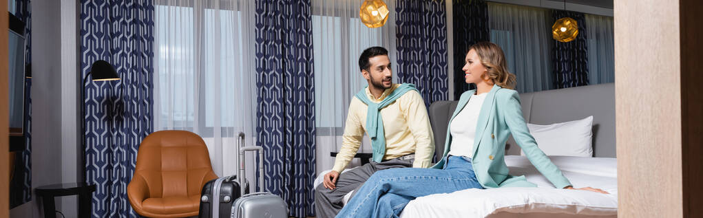 Glückliches interrassisches Paar sitzt auf Hotelbett in der Nähe von Koffern, Banner  - Foto, Bild