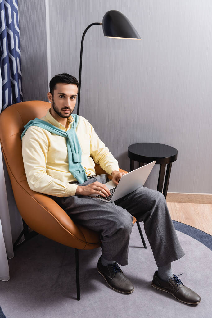 Hombre árabe mirando a la cámara mientras usa el ordenador portátil en el hotel  - Foto, Imagen