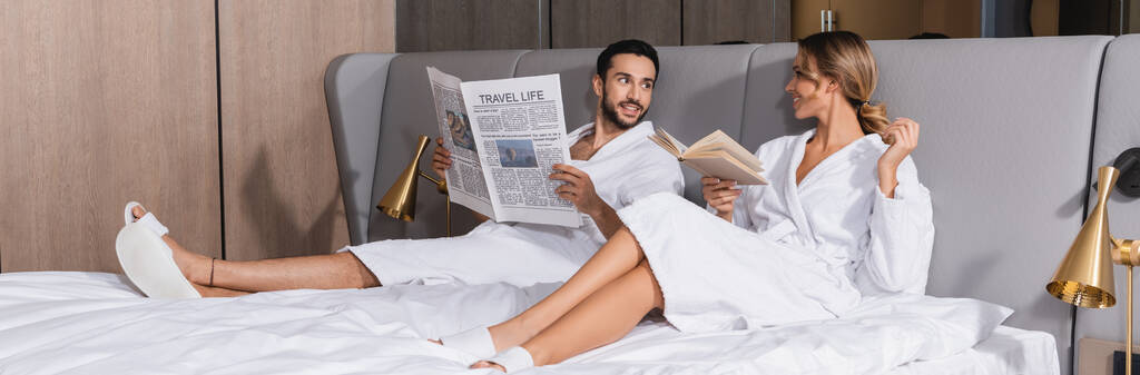 Sonriente pareja interracial con periódico y libro sentado en la cama del hotel, pancarta  - Foto, imagen
