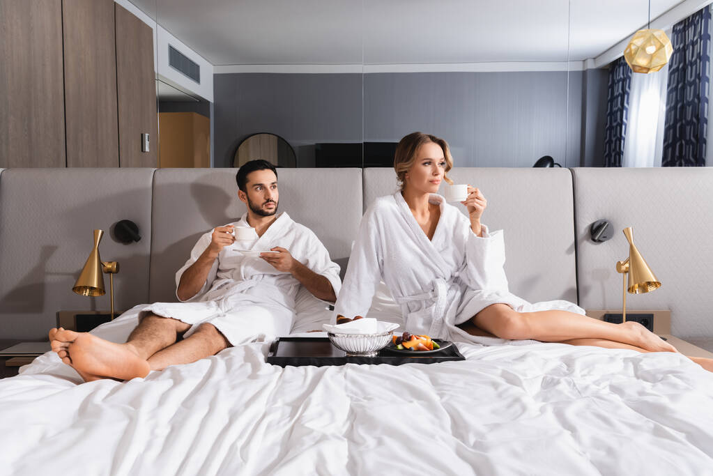 Interracial couple with cups sitting near breakfast on hotel bed  - Photo, Image