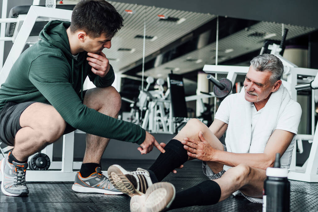 На фото два человека в спортзале, один из них на полу с травмой ноги, тренер по реабилитации помогает ему. - Фото, изображение